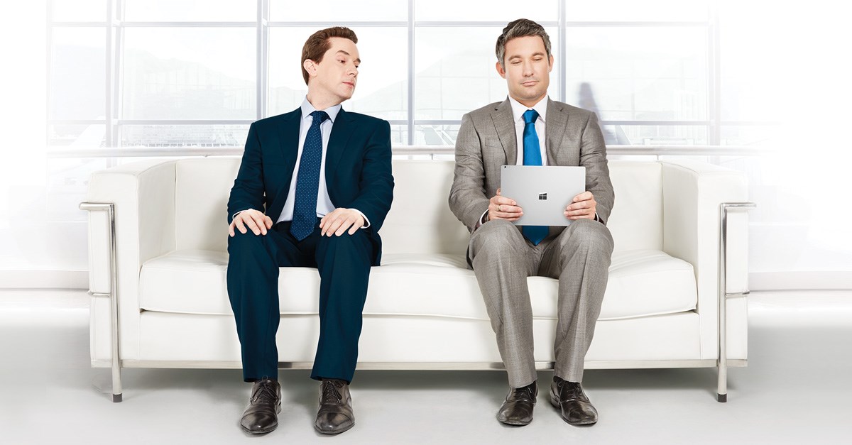 One man using a tablet while another man tries to see what he is doing on the tablet