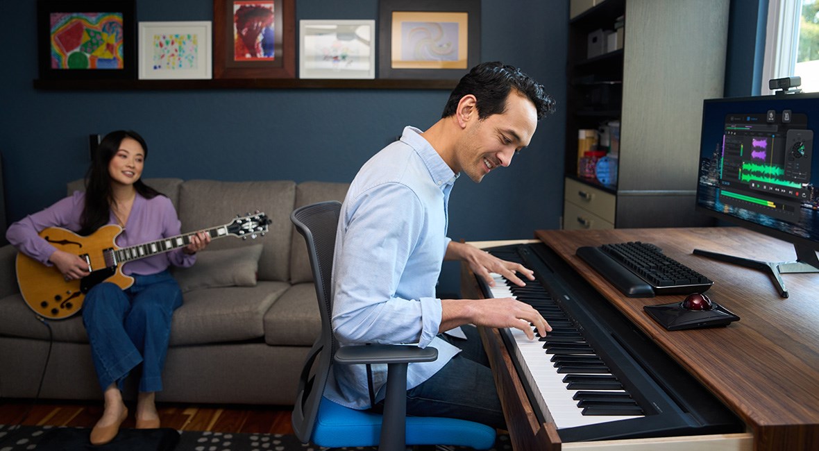 Woman playing guitar while a man in the foreground plays piano at a desk that has a trackball and silent mechanical keyboard and 2 monitors on it.